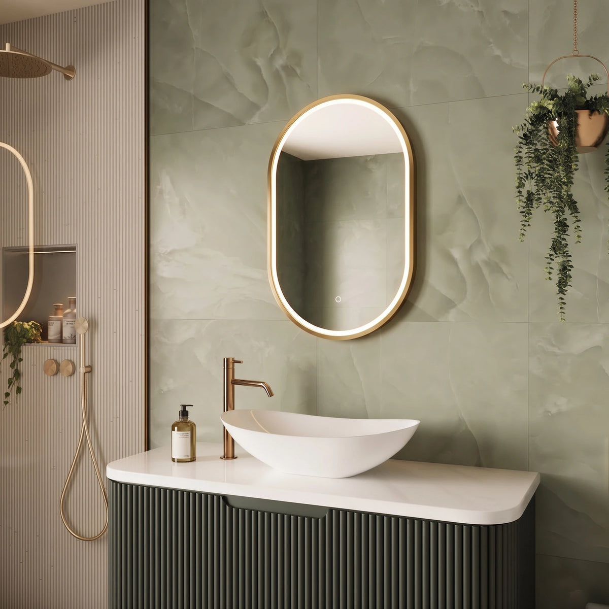A modern bathroom scene featuring an oval backlit LED mirror with a gold frame mounted on a textured sage green tile wall above a white vessel sink and a ribbed dark green vanity cabinet. #Finish_Brushed Brass