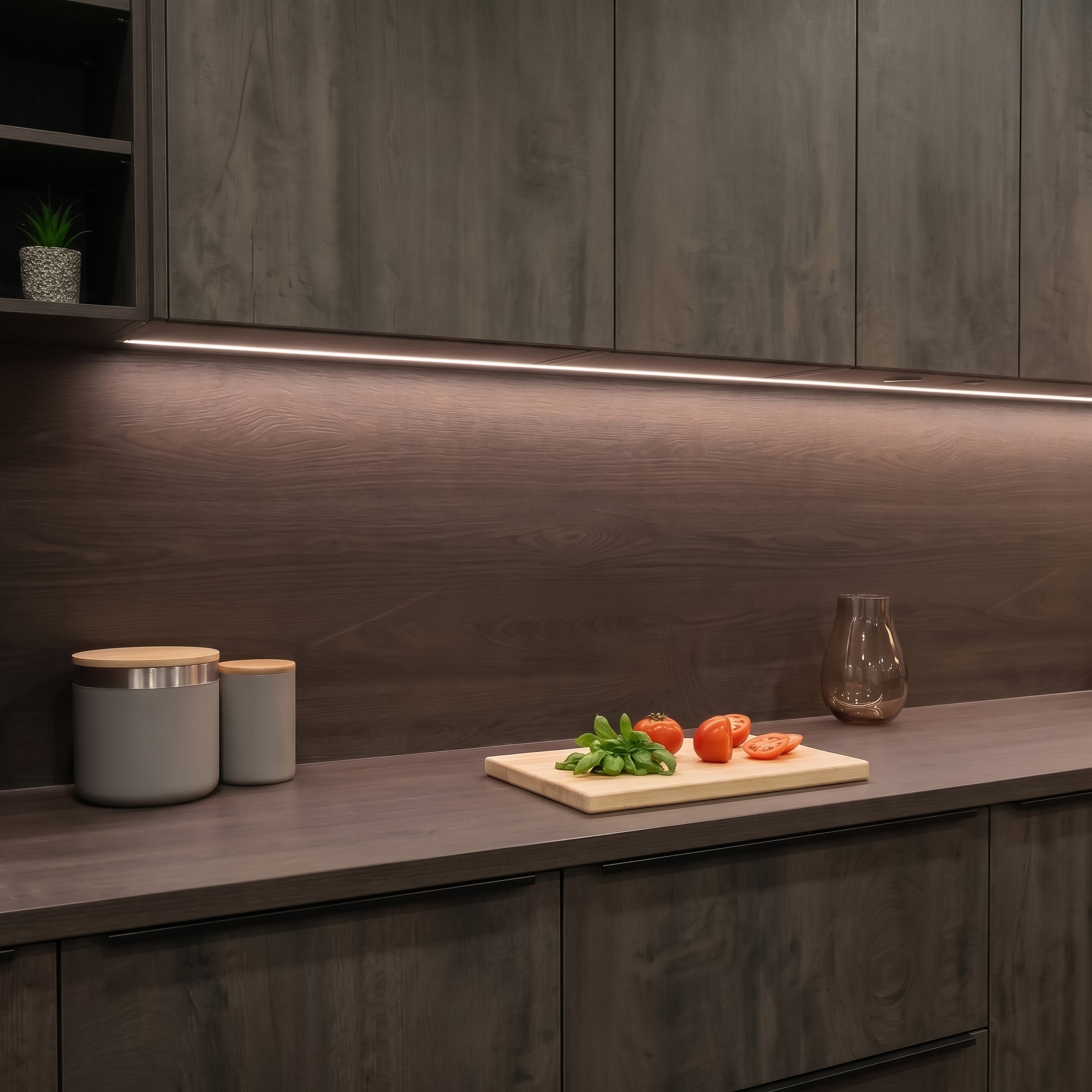 A modern kitchen scene with dark wood cabinets, a matching countertop, and a dark wood backsplash illuminated by under-cabinet LED lighting. A small cutting board with tomatoes and basil sits on the counter, along with two gray canisters and a glass vase. #Finish_Aluminium